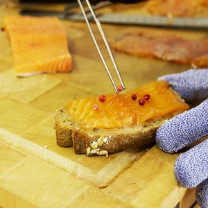 Dressage minutieux d'une tartine de saumon et dépose d'un grain de poivre à l'aide de la pince à dresser